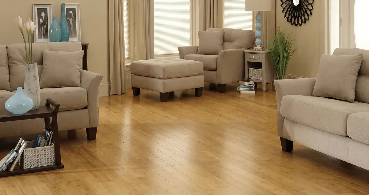 A living room with professionally installed bamboo flooring, beige sofas, a matching ottoman, a side table with a blue lamp, vases, framed pictures, and large windows with blinds and curtains in Myrtle Beach, SC